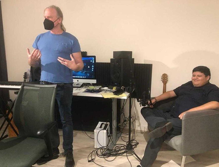 A white man with long gray hair wearing a mask stands speaking with a computer and audio equipment behind him. A Latino teenager wearing black sits in an armchair nearby smiling. Latino Civic Alliance Musician and producer Joe Reineke (left) taught the La